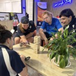 Students enjoying a game of Jenga during a study break at CUC Macleay Valley. Four students sitting around a small table in a study space, playing a game of Jenga together. They are smiling and engaged, with study desks, awards, and a neon ‘Live Laugh Study’ sign visible in the background.