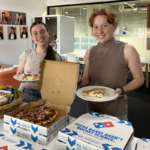 Students take a break to enjoy a shared pizza lunch at CUC Macleay Valley. Two students smiling while serving themselves pizza from open pizza boxes in a communal study space.