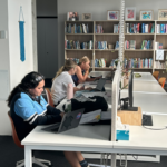 Students concentrating on their work using laptops at a row of study desks, surrounded by resources on a full bookshelf. Three young women studying at white desks in a well-lit modern study space or library with a large bookshelf filled with books in the background.