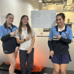 Three students show off their progress on a collaborative, physical Wordle game set up in a common area. Three high school students posing and pointing at a large, hand-drawn Wordle grid on a whiteboard or poster board behind them.